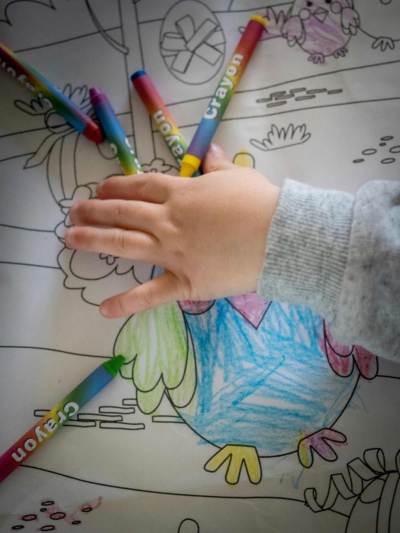 A child's hand resting on a colouring sheet among wax crayons